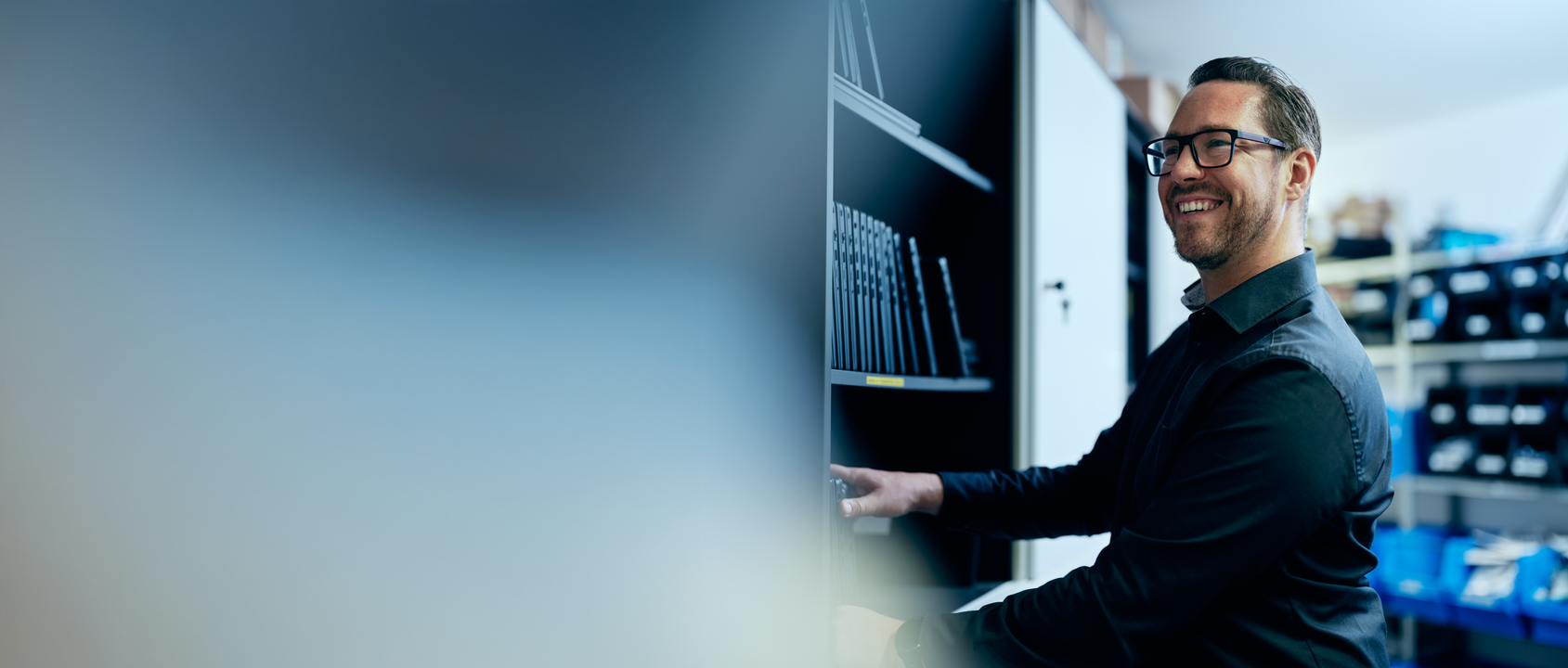 Bild von Ernest Vögerle auf dem er vor einem Schrank mit Laptops steht. Ernest ist zur Seite gedreht und lächelt.