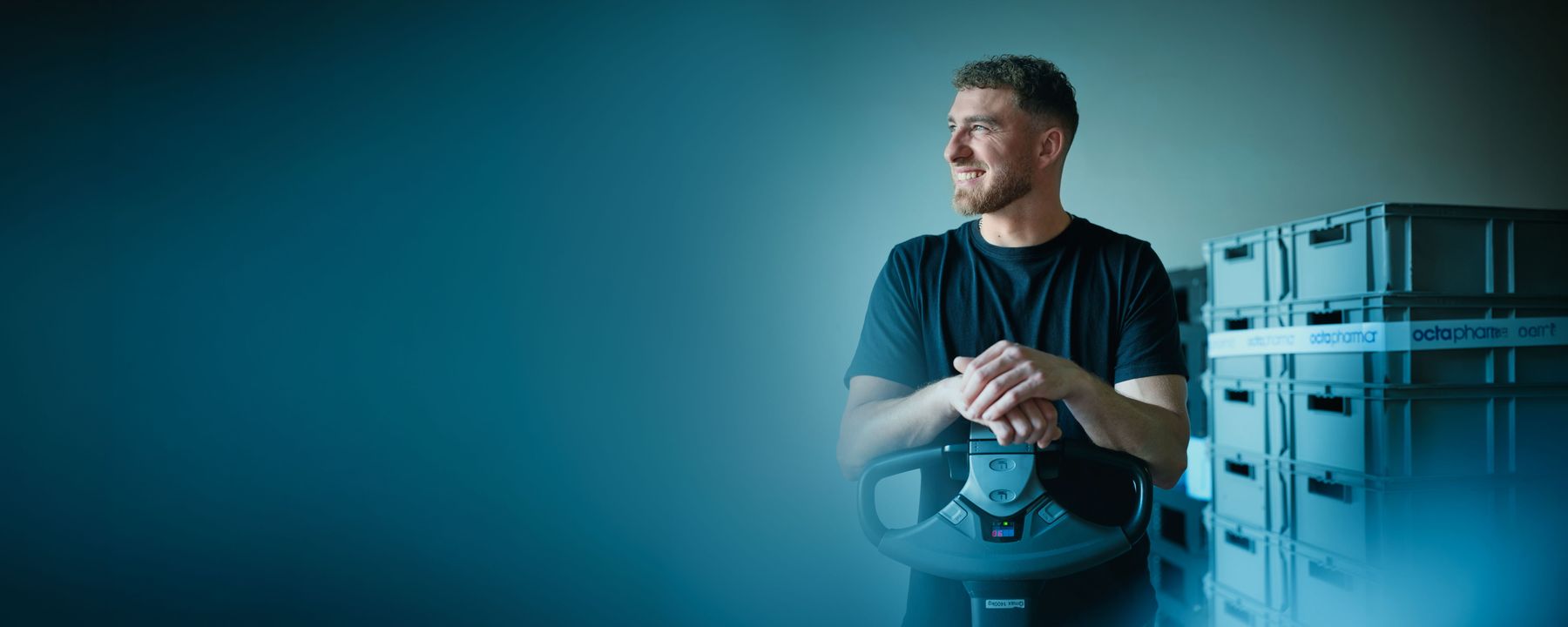 Smiling man in a black t-shirt leans on a steering wheel near stacked storage boxes, with a soft blue gradient background.
