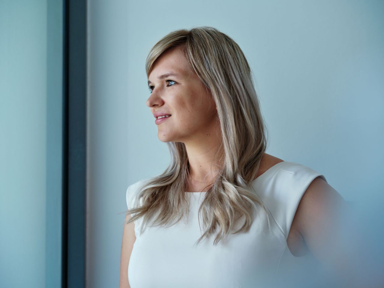 A woman with long blonde hair wearing a white top gazes thoughtfully out of a window, with a soft blue background.