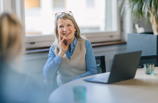Smiling woman with glasses on head, wearing a blue shirt and vest, sits at a table with a laptop, engaging in conversation.