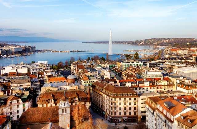 Aerial view of Geneva with the Jet d'Eau fountain, Lake Geneva, and surrounding buildings under a clear blue sky.