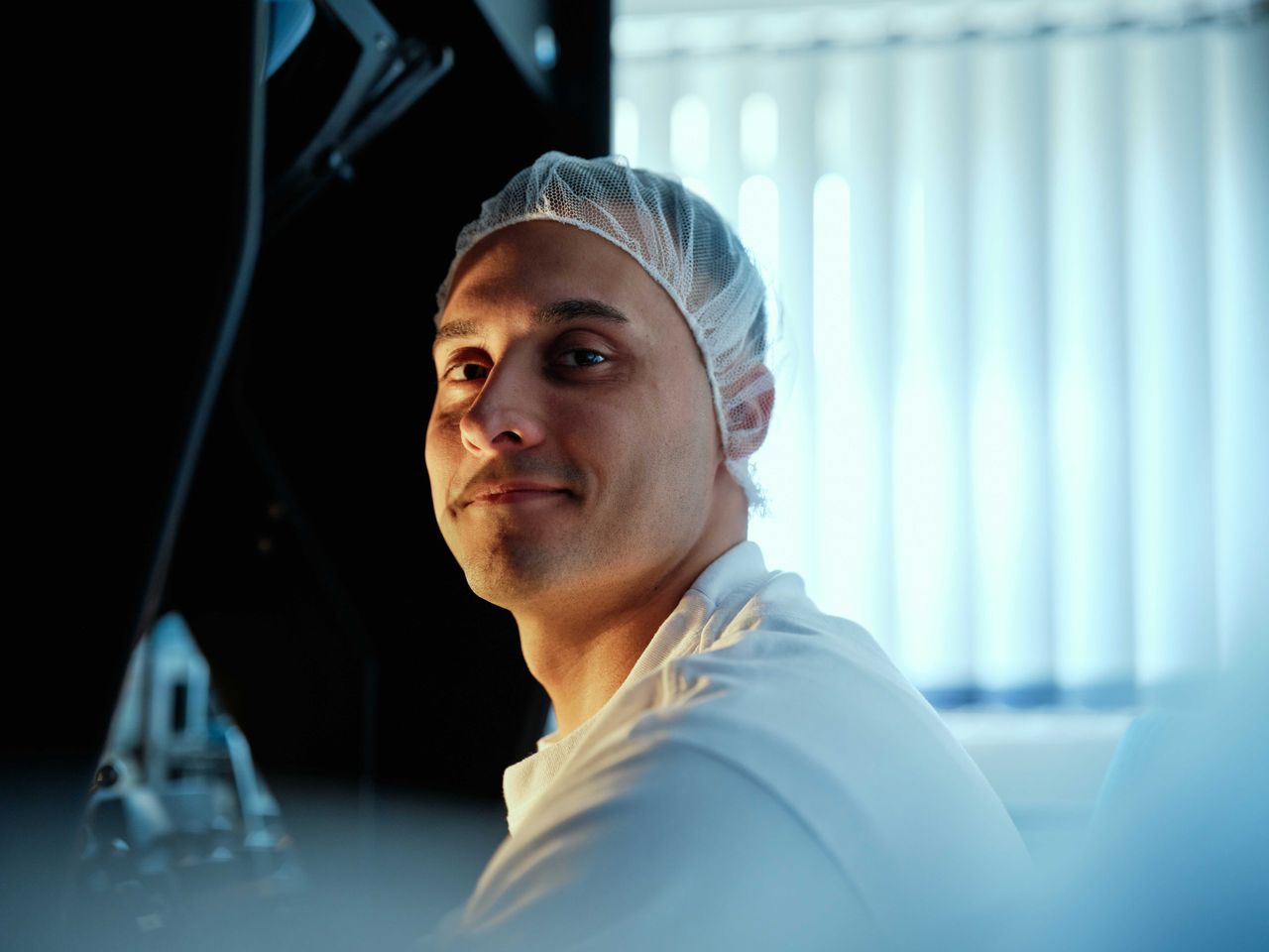 A person wearing a hairnet and white shirt smiles slightly, with vertical blinds and a machine visible in the background.