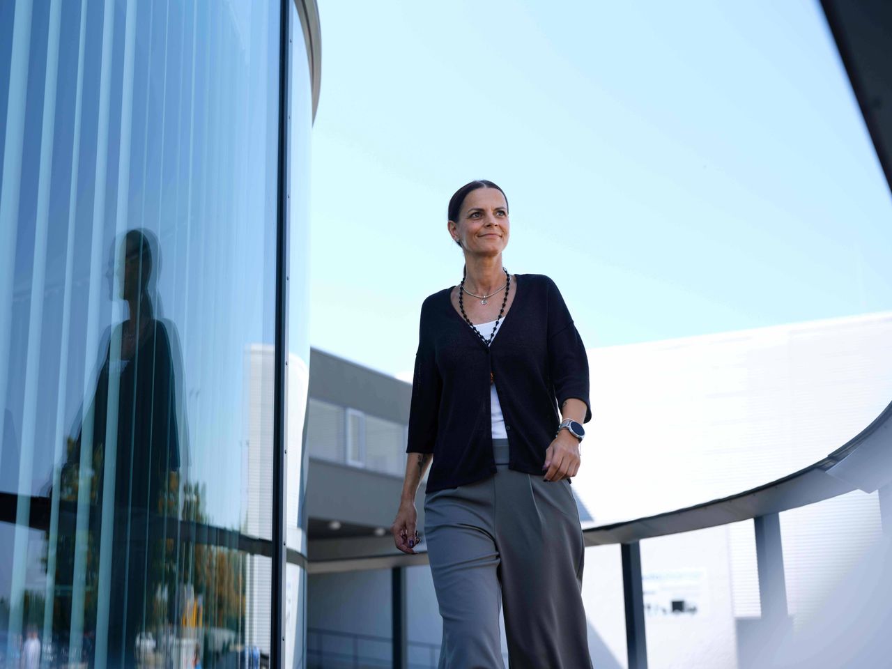 A woman walks confidently past a modern glass building under a clear blue sky, her reflection visible in the glass.