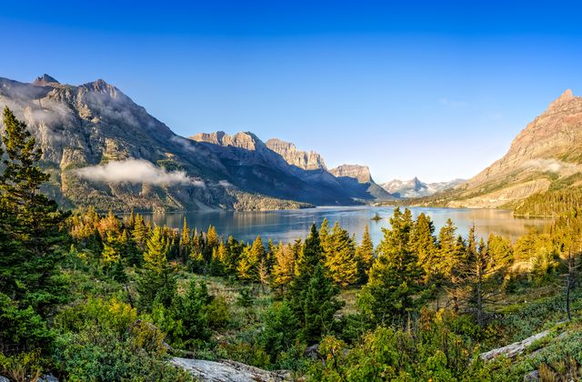 Mountain landscape with a clear blue sky, a serene lake, pine trees in the foreground, and distant peaks partially covered in mist.