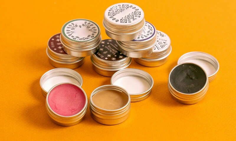 Stacked tins of lip balms in a range of shades and textures on a warm background, representing the modern lip care ritual where fragrance, feel, and emotion combine to create sensorial, feel-good beauty experiences