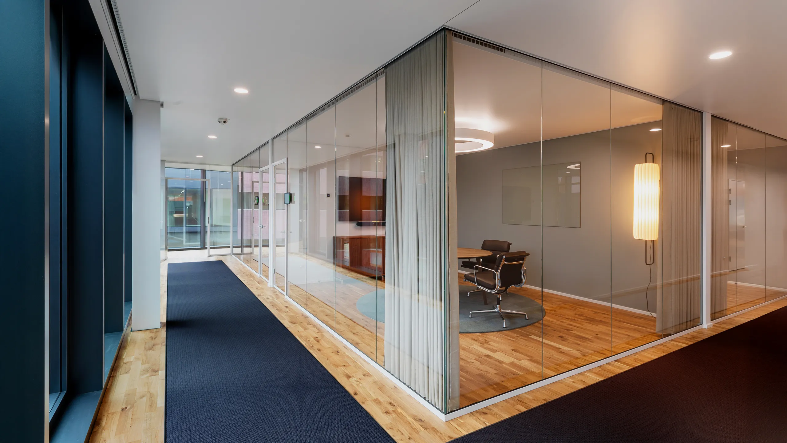 Modern office hallway with glass-walled meeting room, wooden floors, and minimalistic decor. A circular overhead light is visible inside.