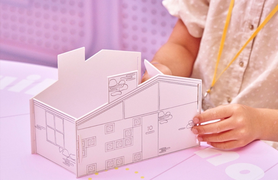 A child builds a paper house on a pink table
