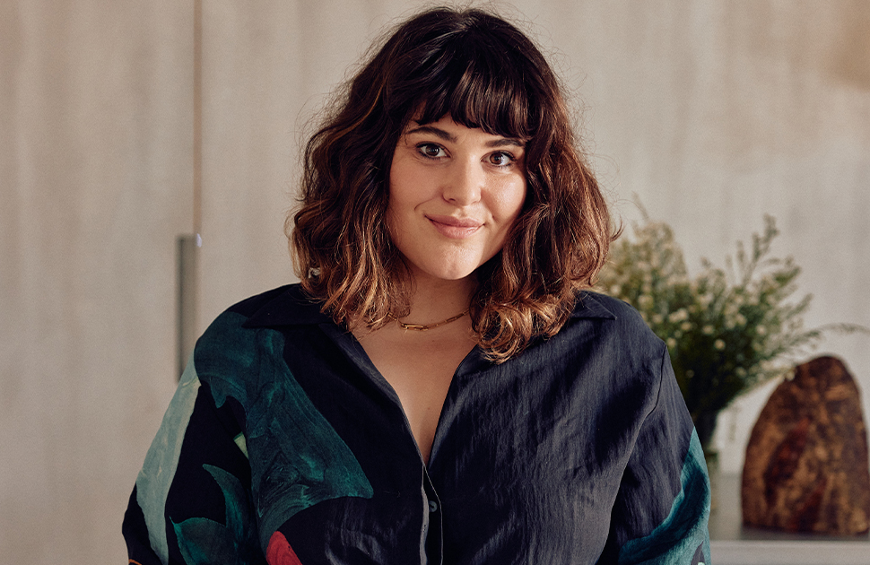 Julia Busuttil Nishimura stands in front of light wooden floor to ceiling cupboards with small white floral bouquet and rock on bench behind her
