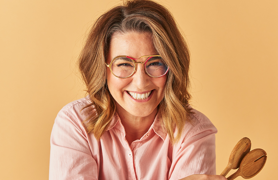 Alice Zazlavsky smiling at the camera, holding salad spoons, in front of an orange background