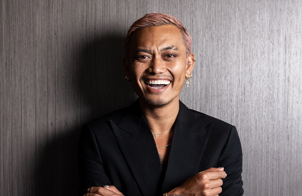 Khanh Ong smiles at camera, standing front of dark, wooden textured wall