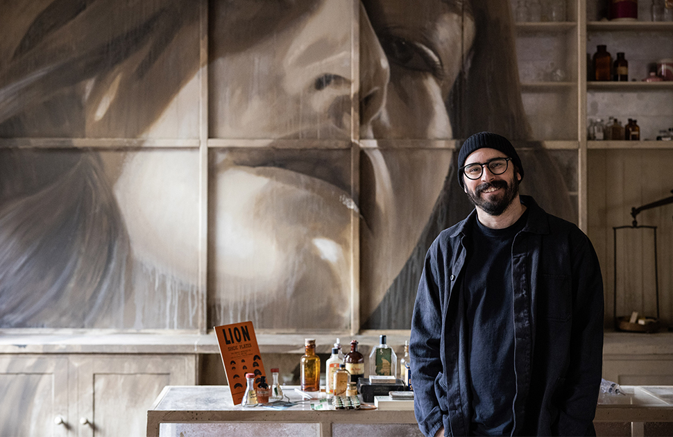 A photo of RONE standing in front of a mural of a woman's face he painted on a wall. He leans against a table with various bottles on it.