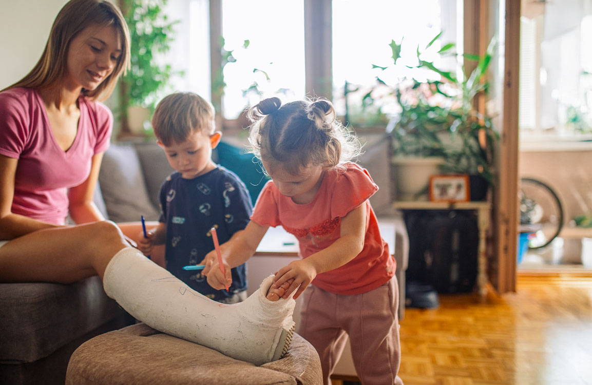 Mutter mit gebrochenem Bein sitzt auf Sofa und lässt ihre Kinder den Gipsverband bemalen