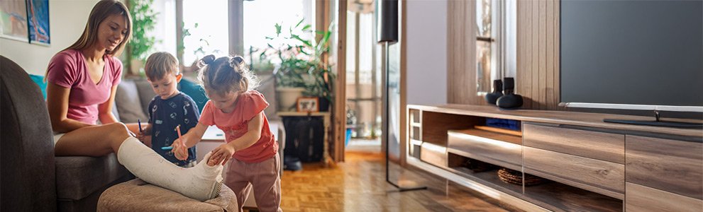 Mutter mit gebrochenem Bein sitzt auf Sofa und lässt ihre Kinder den Gipsverband bemalen