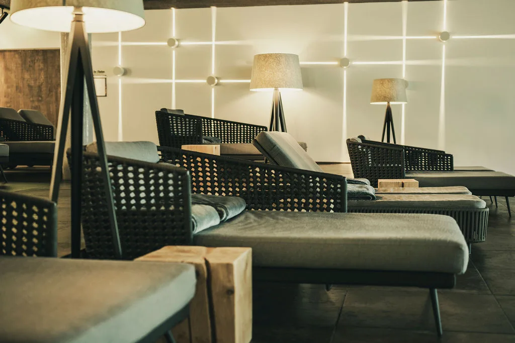 Modern lounge with wicker chaise lounges, cushioned seats, wooden side tables and tripod floor lamps under soft wall lighting.