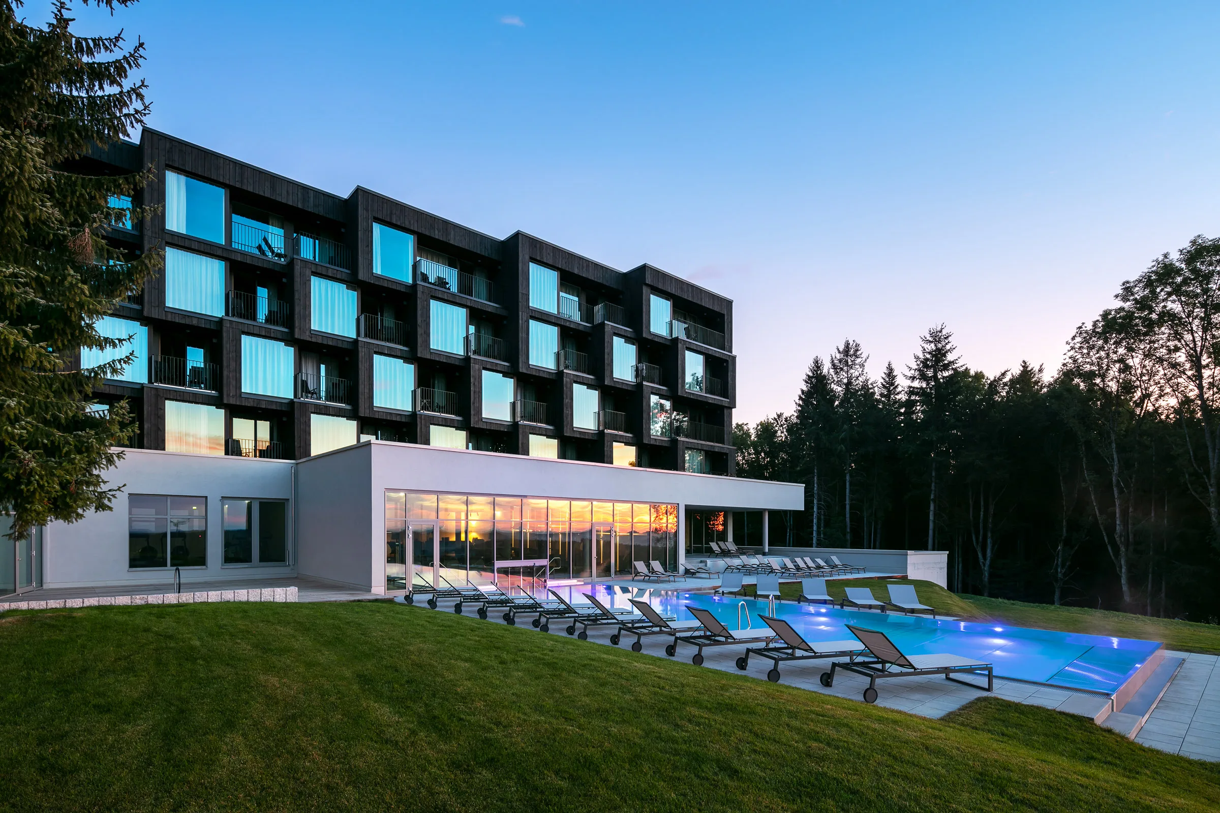 Modern hotel with a sleek design, large windows, and a pool surrounded by lounge chairs, set against a backdrop of trees at sunset.