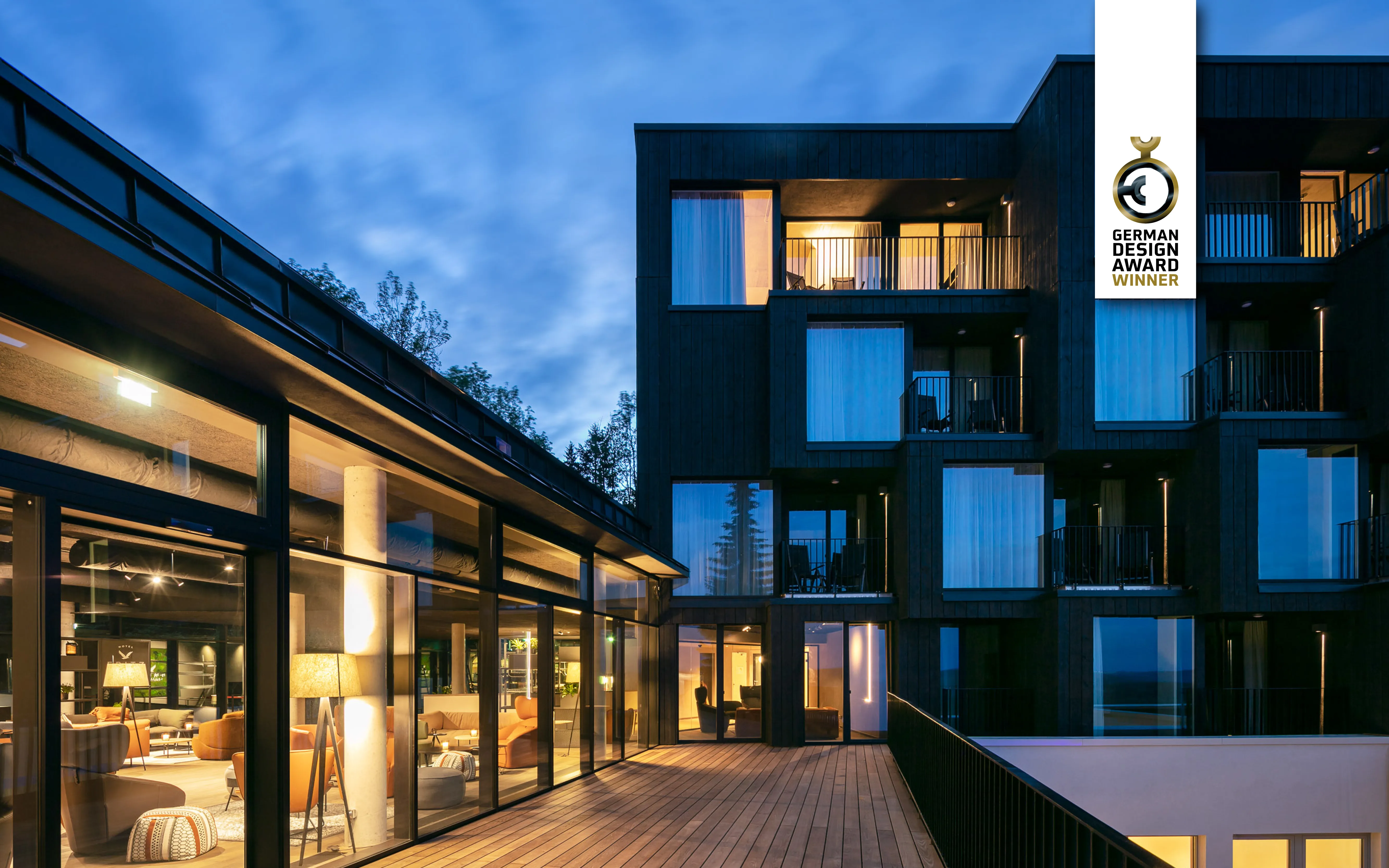 Modern building with large windows and a wooden deck at dusk, featuring a "German Design Award Winner" badge.
