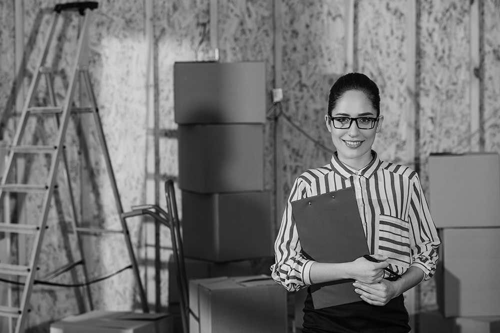 A businesswoman in wearing glasses and a holding a clipboard smiles in front of inventory boxes in a newly constructed room with bare wood framed walls.