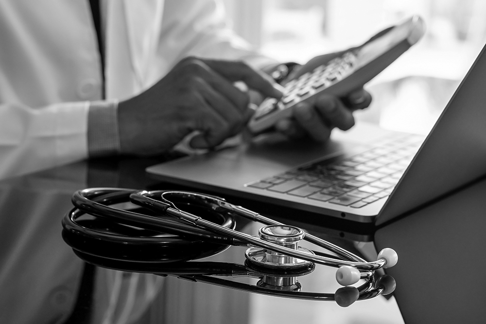 A medical professional in a white lab coat with a stethoscope on the desk uses a pocket calculator. 