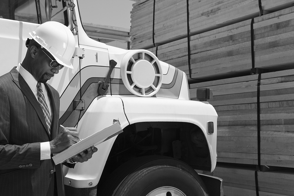 A businessman in a hardhat standing next to his new truck as he signs on a clipboard.