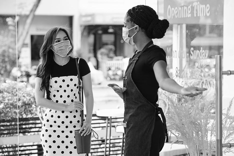 A face mask wearing shop owner opens her dorr and welcomes a mask-wearing customer into her business.