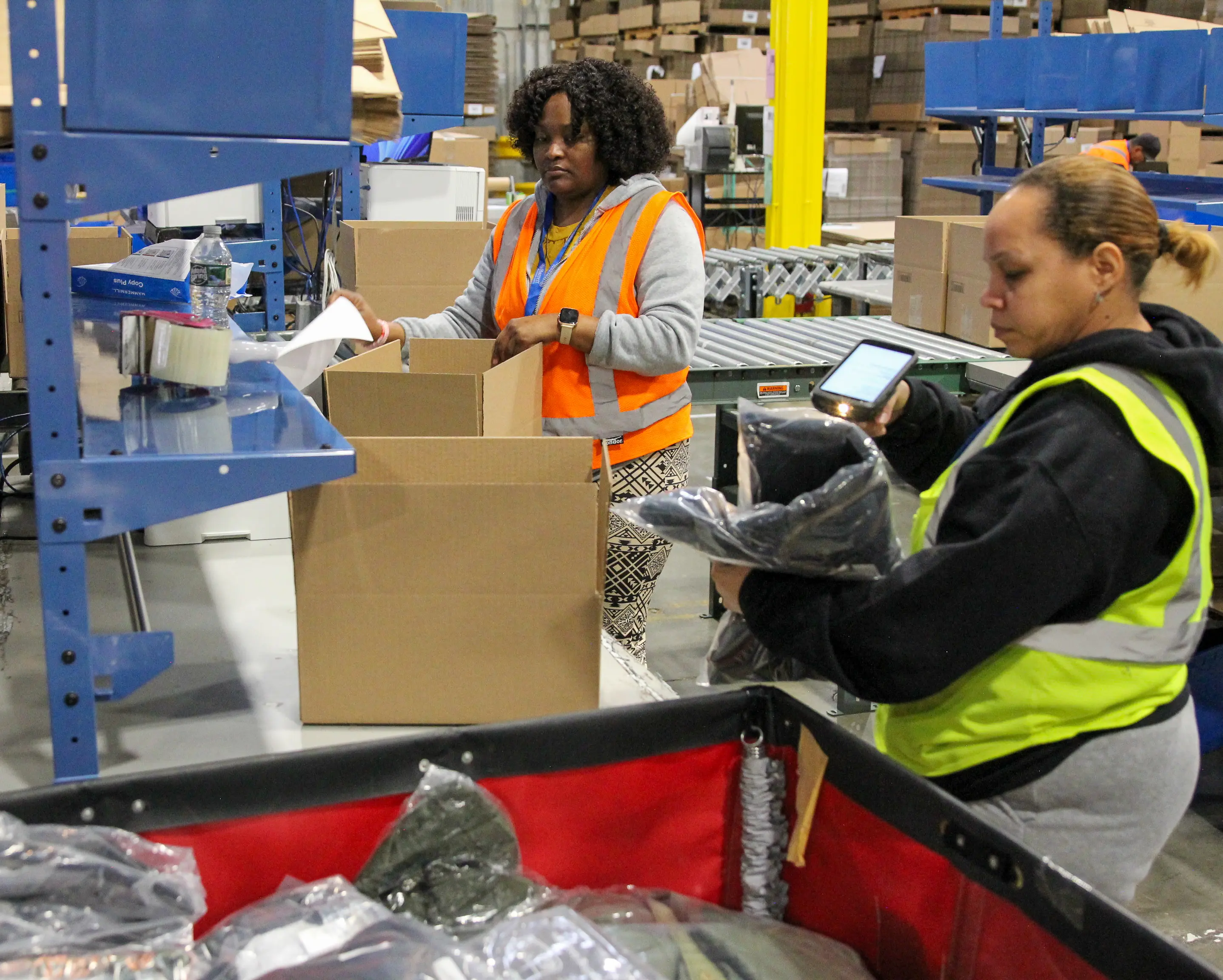 Two workers in a warehouse, wearing safety vests, are packing and scanning items into boxes, surrounded by shelves and bins at a pack station.