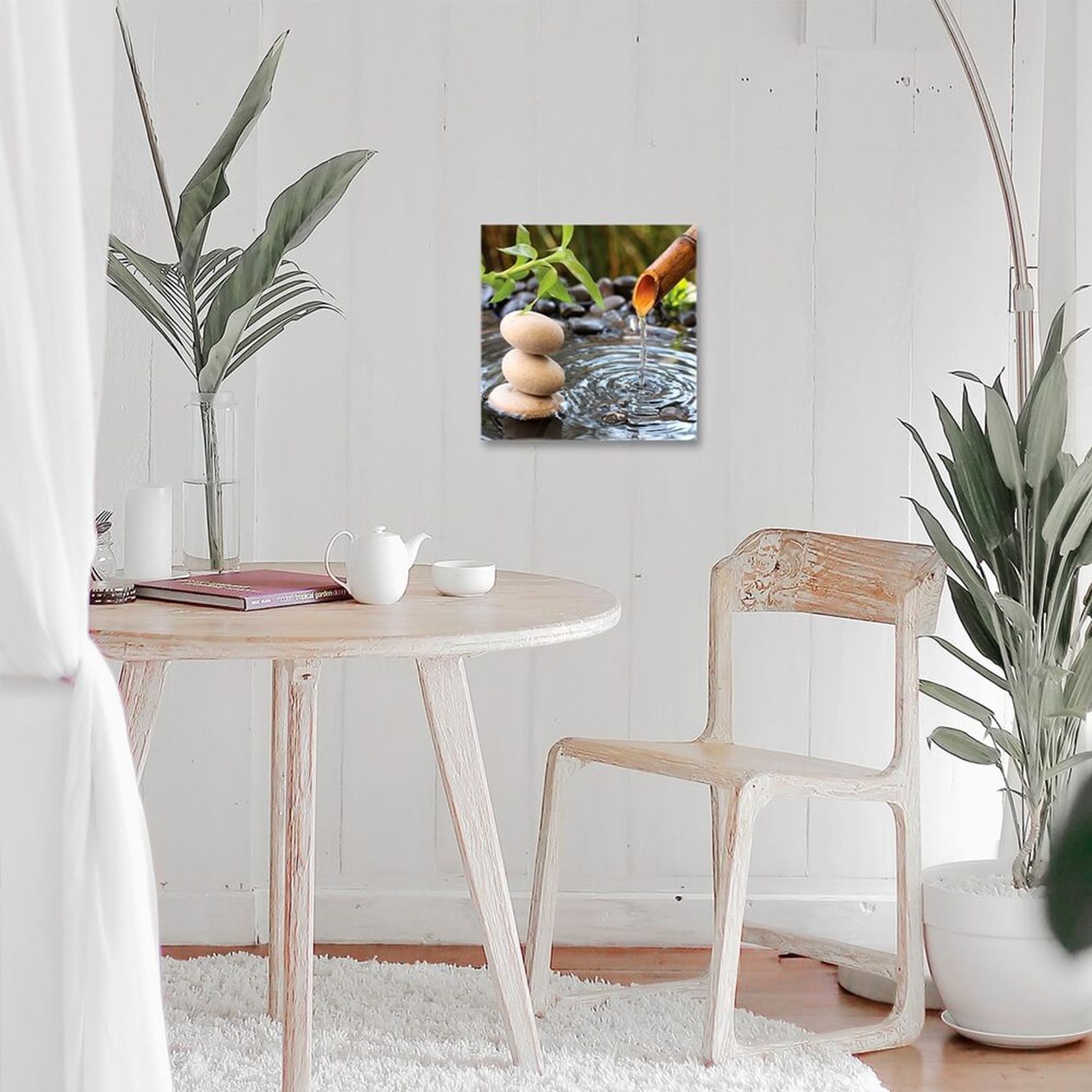 Quadratisches Glasbild mit Zen-Motiv – gestapelte Steine, Bambusbrunnen und fließendes Wasser – an einer weißen Holzwand über einem kleinen Tisch mit Teekanne.