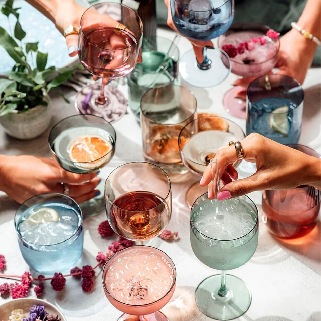 Mehrere bunte Wein- und Cocktailgläser in Rosa, Blau, Grün und Lila werden von Händen angestoßen; auf dem Tisch Beeren, Zitrusfrüchte und getrocknete Blumen.