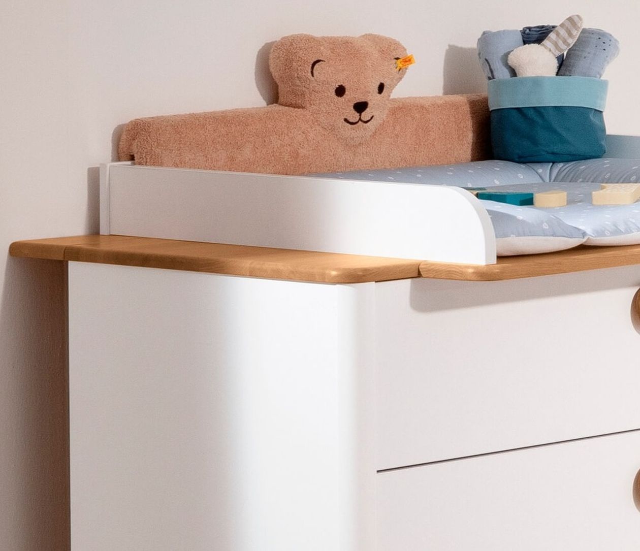 Nahaufnahme eines weißen Wickelaufsatzes auf einer Holzkommode mit einem Teddybär und einem blauen Stoffkorb auf der Wickelunterlage.