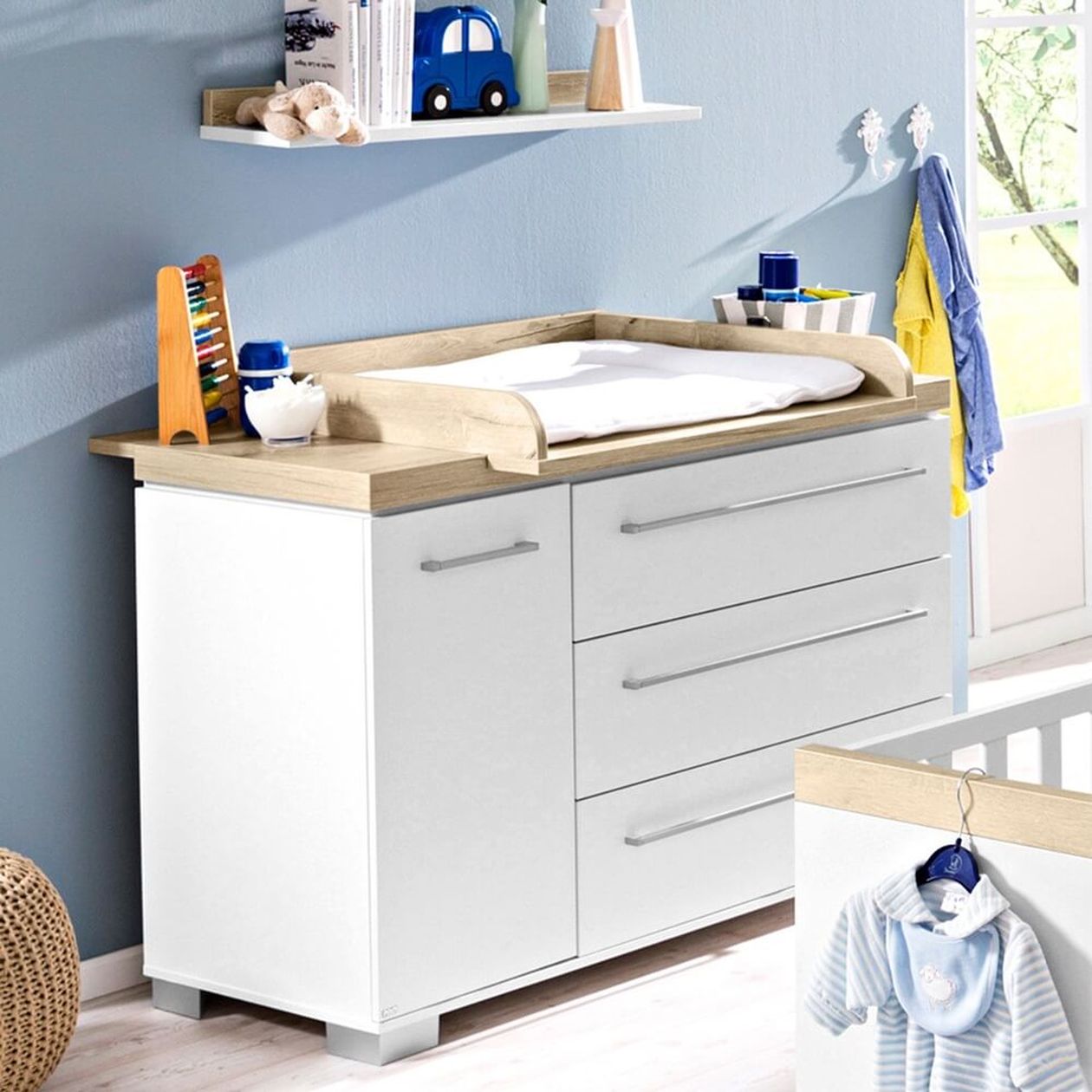 Weiße Wickelkommode mit naturholzfarbener Platte und Wickelaufsatz mit hellblauer Unterlage, drei Schubladen und einer Tür, eingerichtet in einem hellblauen Babyzimmer.