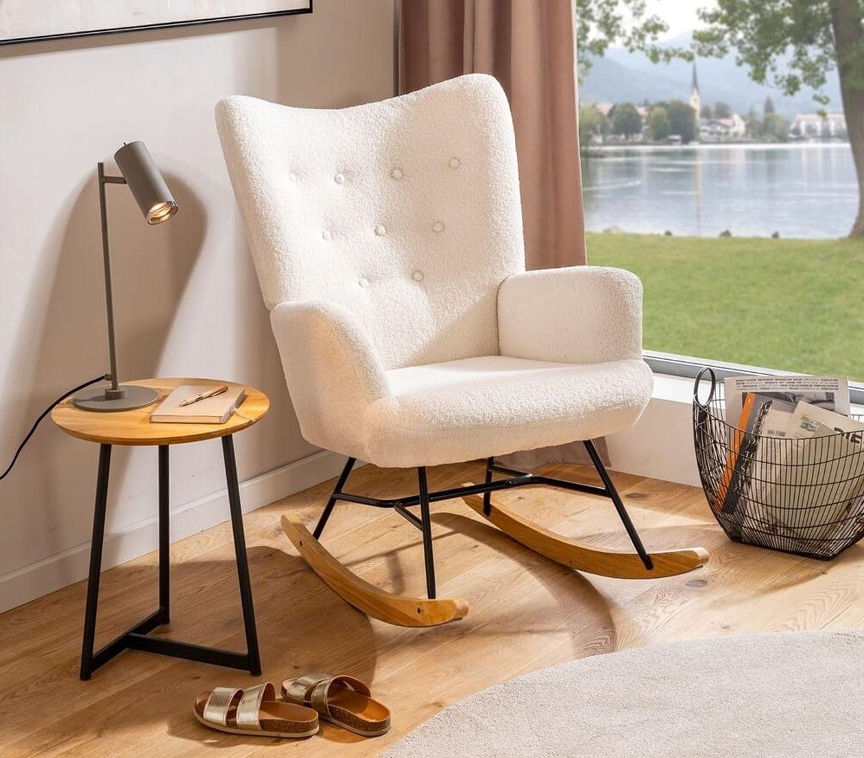 Cremefarbener Schaukelstuhl mit Bouclé-Bezug und Holzkufen vor einem großen Fenster mit Blick auf einen See, daneben ein Beistelltisch mit Lampe und ein Drahtkorb.