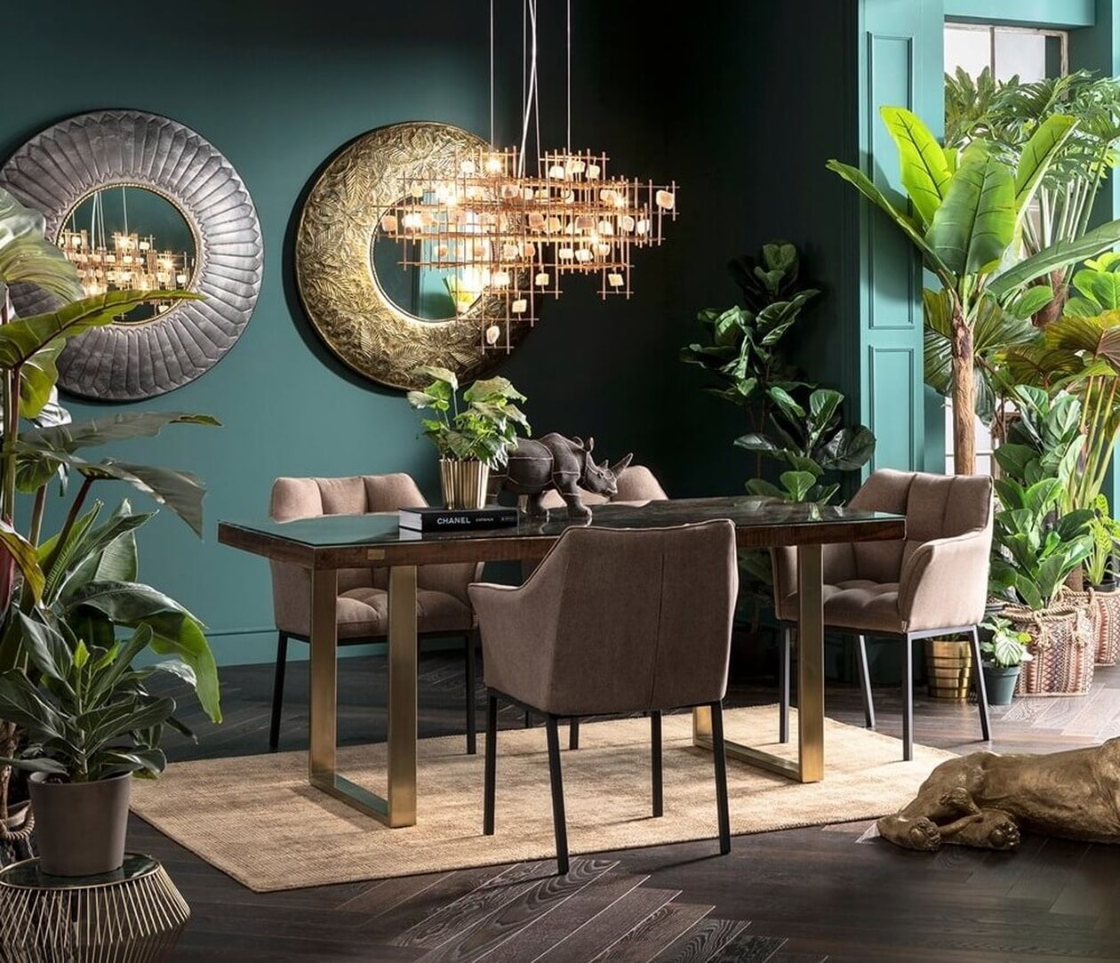 Elegantes Esszimmer mit dunkelgrünen Wänden, einem Esstisch mit Glasplatte und goldenen Beinen, umgeben von beigefarbenen Polsterstühlen. Dekorative Wandspiegel, eine goldene Hängeleuchte und zahlreiche große Grünpflanzen, darunter eine Bananenpflanze, verleihen dem Raum tropisches Flair.