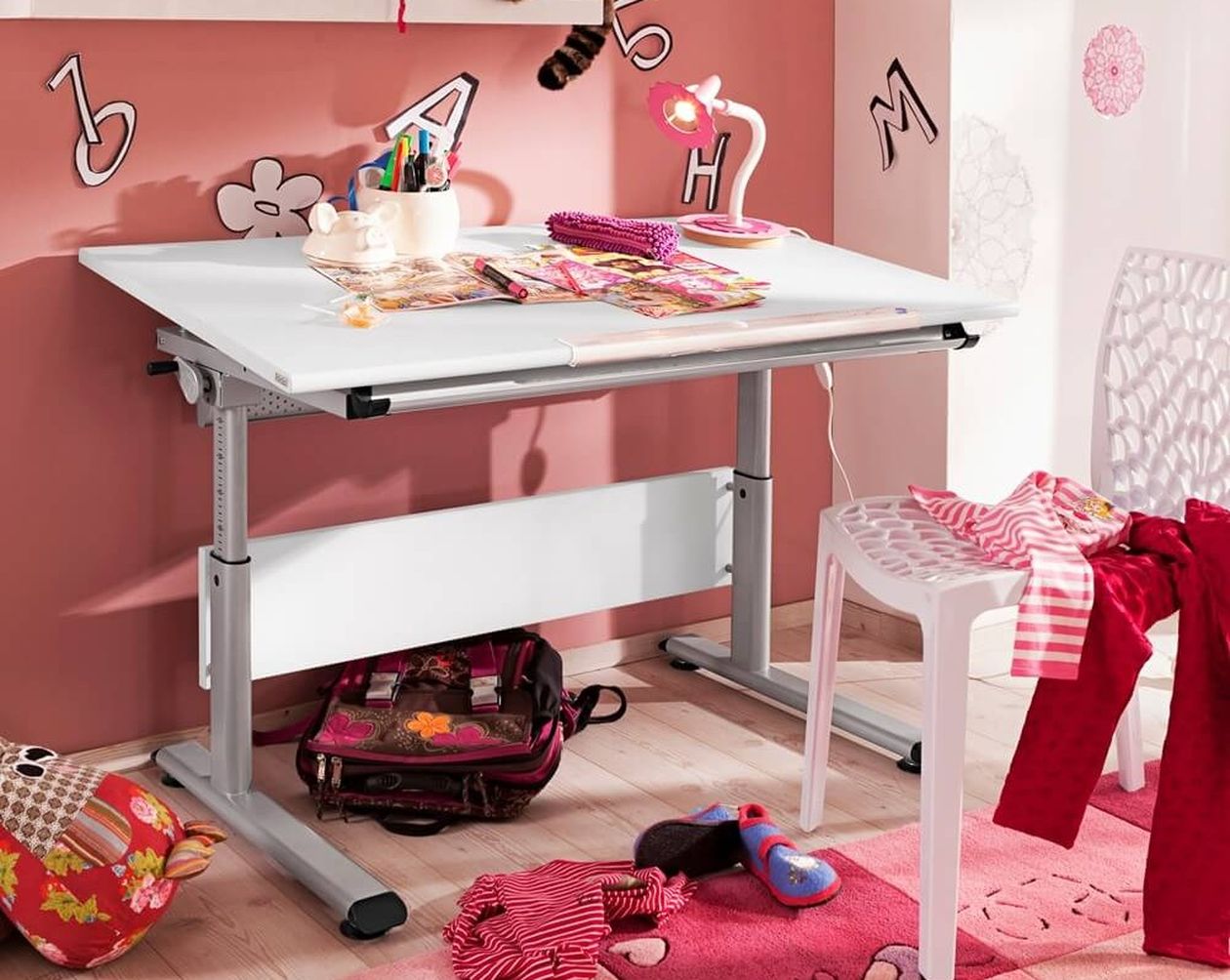 Höhenverstellbarer weißer Kinderschreibtisch mit neigbarer Tischplatte und silbernem Metallgestell in einem rosa Kinderzimmer. Auf dem Tisch Bastelmaterial und Zeitschriften, darunter ein bunter Schulranzen.