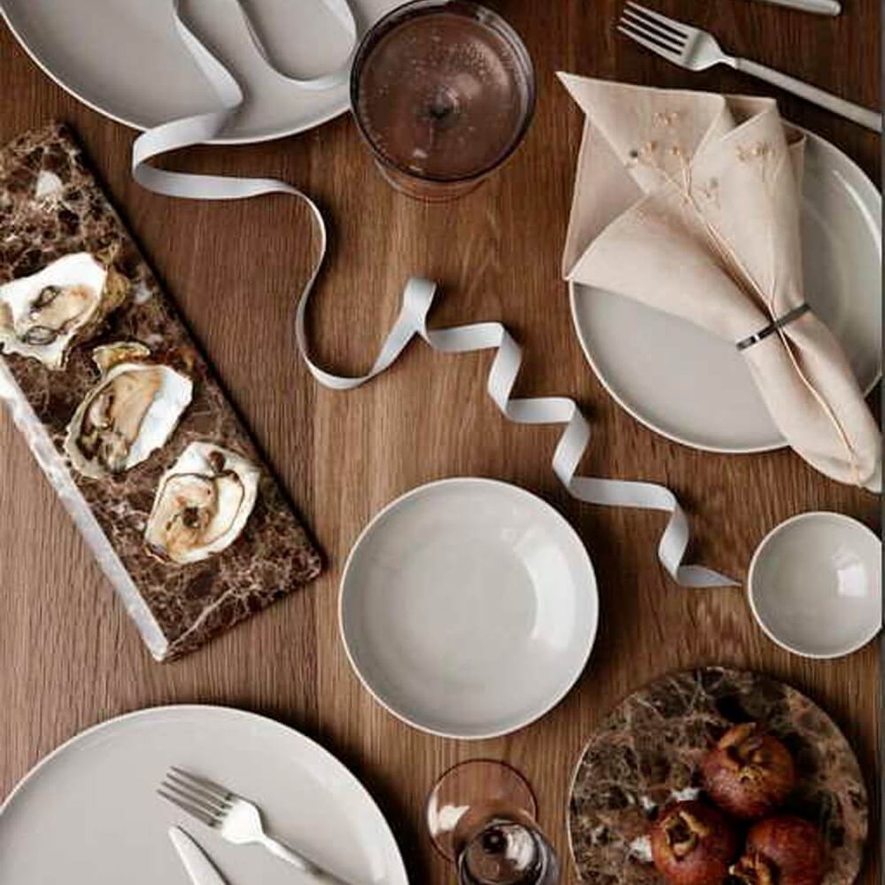 Gedeckter Holztisch von oben mit hellem Kombiservice aus Tellern, Schalen und Gläsern in Grau-Beige, Austern auf Marmorplatte, beiger Leinserviette und dekorativem weißem Band.