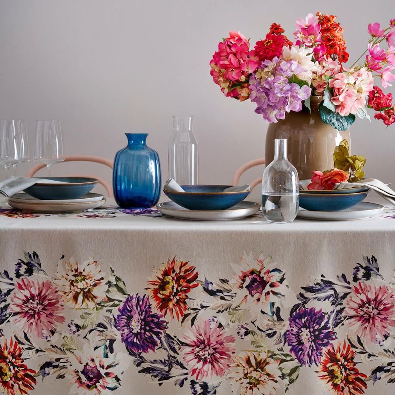 Gedeckter Tisch mit floralem Tischläufer in kräftigen Rosa-, Rot- und Lilatönen, blauem und klarem Glasgeschirr sowie einem üppigen Blumenstrauß in brauner Vase.