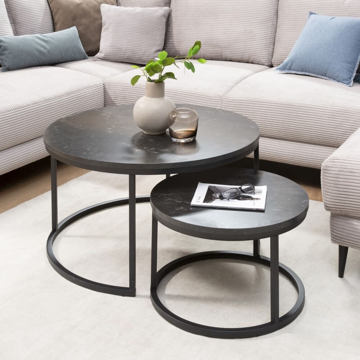 Zwei runde, ineinandergreifende schwarze Couchtische mit einer Vase, einer Pflanze, einer Kerze und einem Magazin darauf, in einem gemütlichen Wohnzimmer mit einem beigen Ecksofa.