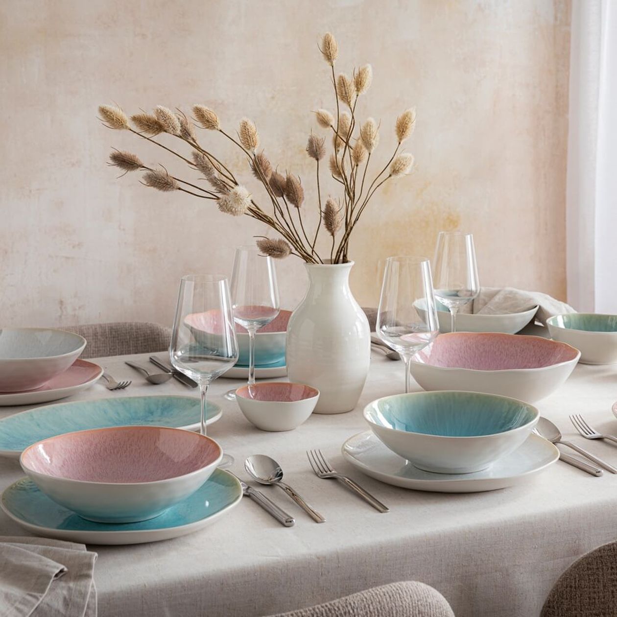 Gedeckter runder Tisch mit rosa-blauen Keramikschalen in Farbverlauf-Glasur, Weingläsern und weißer Vase mit Trockenblumen auf hellem Leinentischtuch.