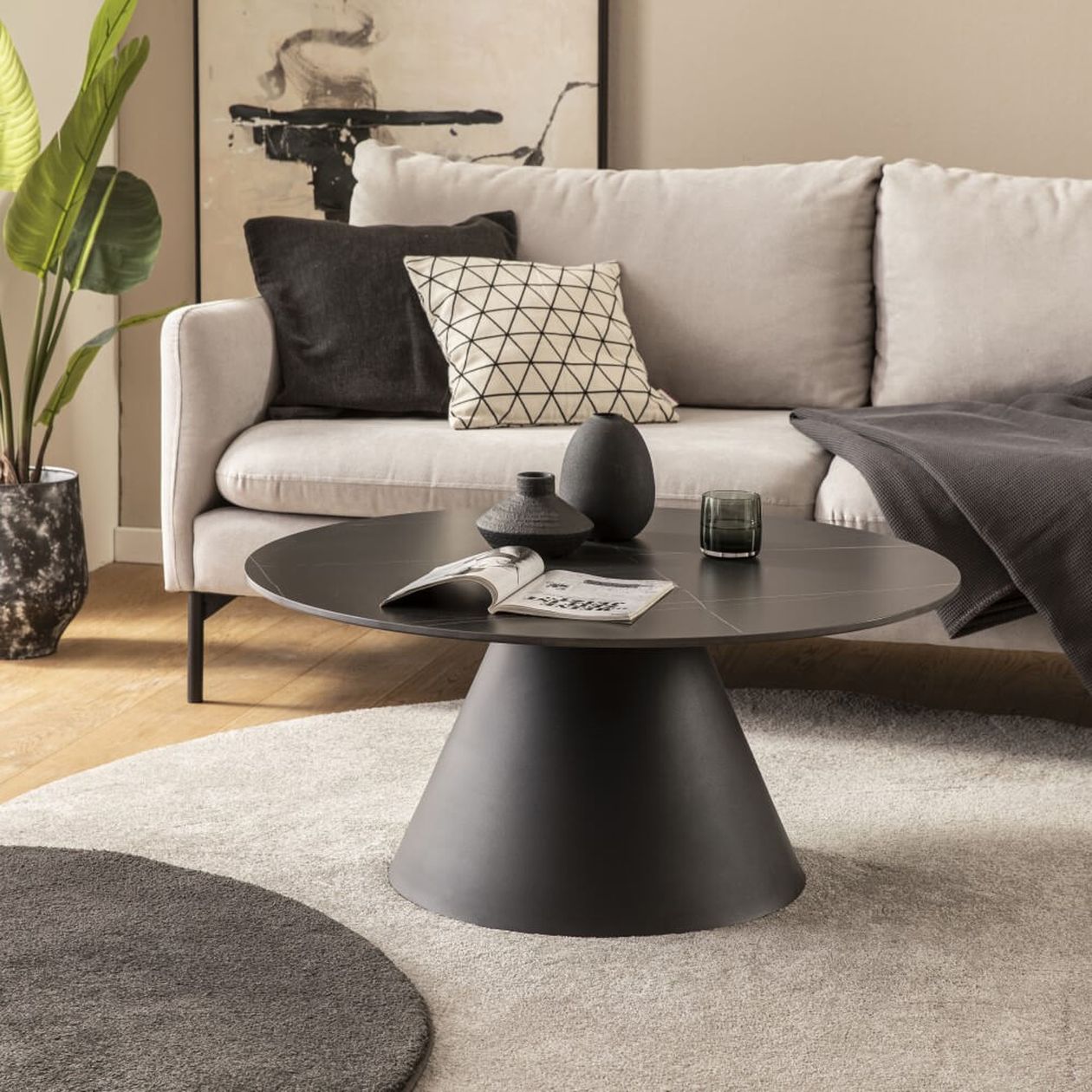 Modernes Wohnzimmer mit einem beigen Sofa, geometrischen Kissen, schwarzem runden Couchtisch und Dekorationsartikeln. Große Pflanze und abstrakte Kunst im Hintergrund.