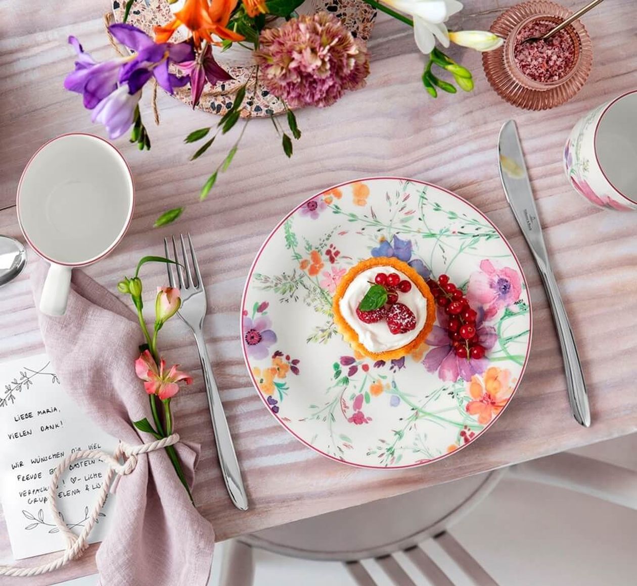 Gedeckter Platz von oben: Frühstücksteller mit buntem Blumenmuster und einem Törtchen mit Beeren, flankiert von Besteck und lila Serviette mit Blume; im Hintergrund Blumenstrauß und Tassen.