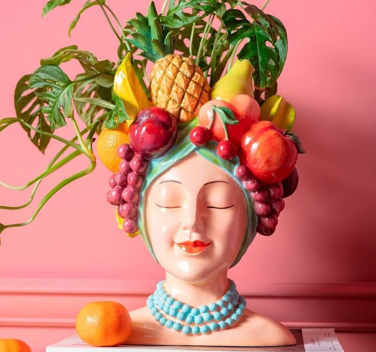 Bunte Keramikvase in Form eines Frauenkopfes mit Früchten als Kopfschmuck im Carmen-Miranda-Stil. Grüne Pflanze als Inhalt, vor einer pinken Wand auf Büchern platziert.