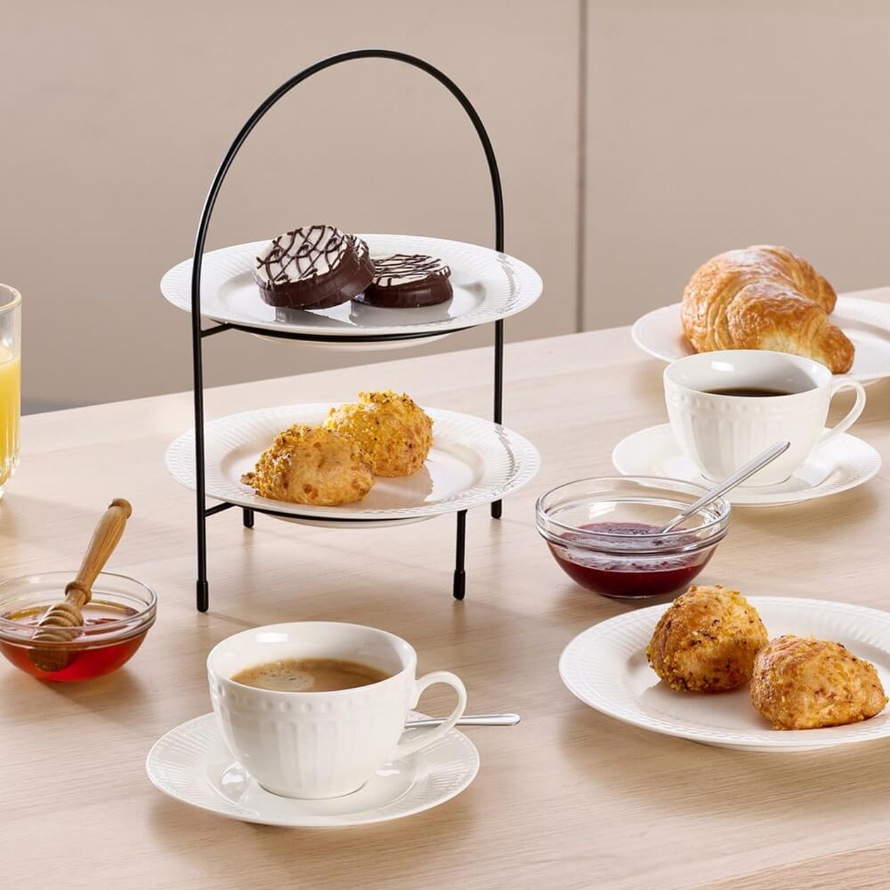 Zweistöckige Etagere mit schwarzem Metallgestell und weißen Keramiktellern, beladen mit Schokoladenplätzchen und Gebäck, daneben Kaffeetassen, Croissants, Marmelade und Honig auf hellem Holztisch.