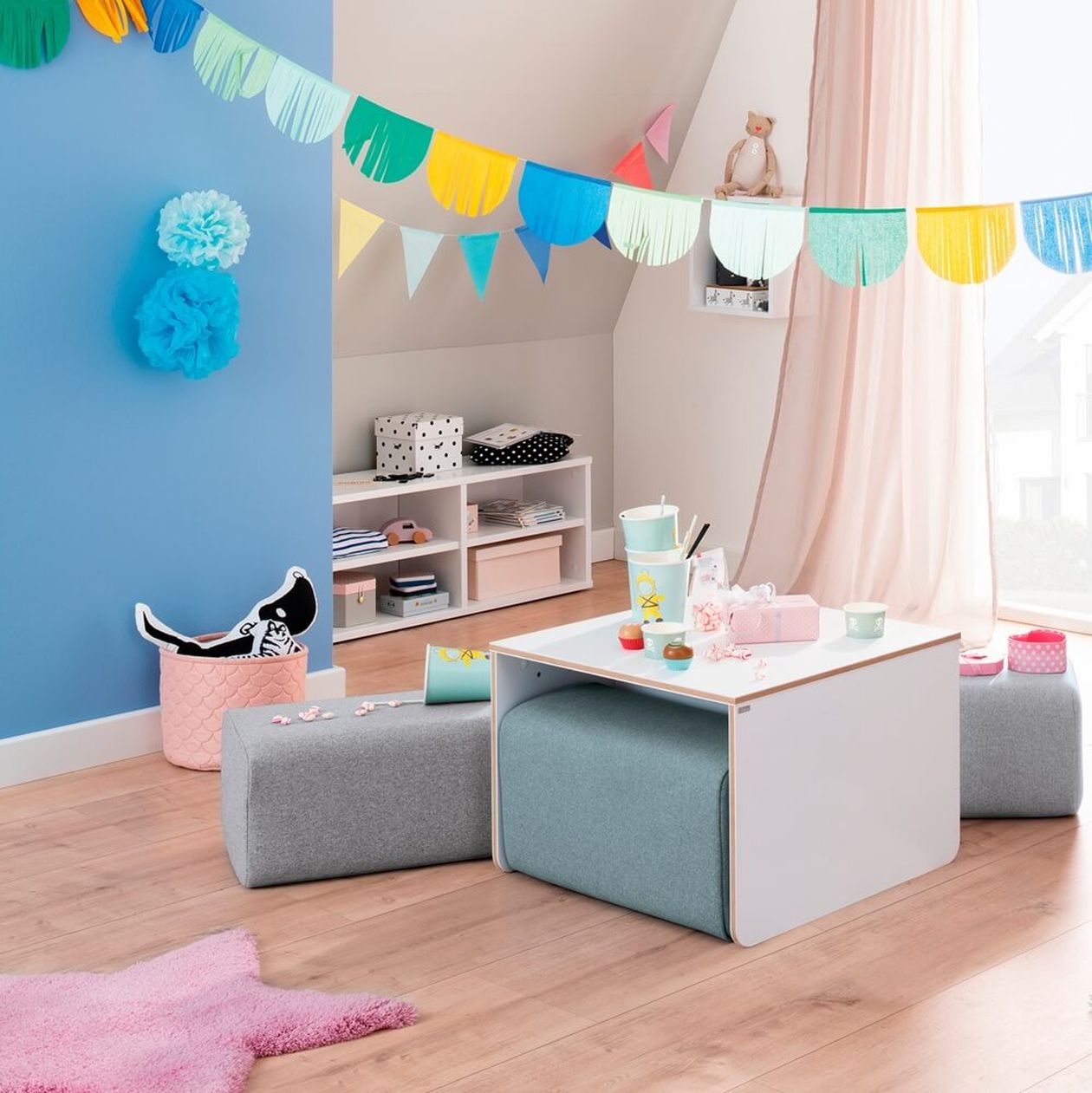 Kinderzimmer mit hellblauer Wand, bunte Wimpelkette an der Decke und Sitzwürfeln um einen weißen Tisch. Im Hintergrund ein kleines Regal und Dekoration.