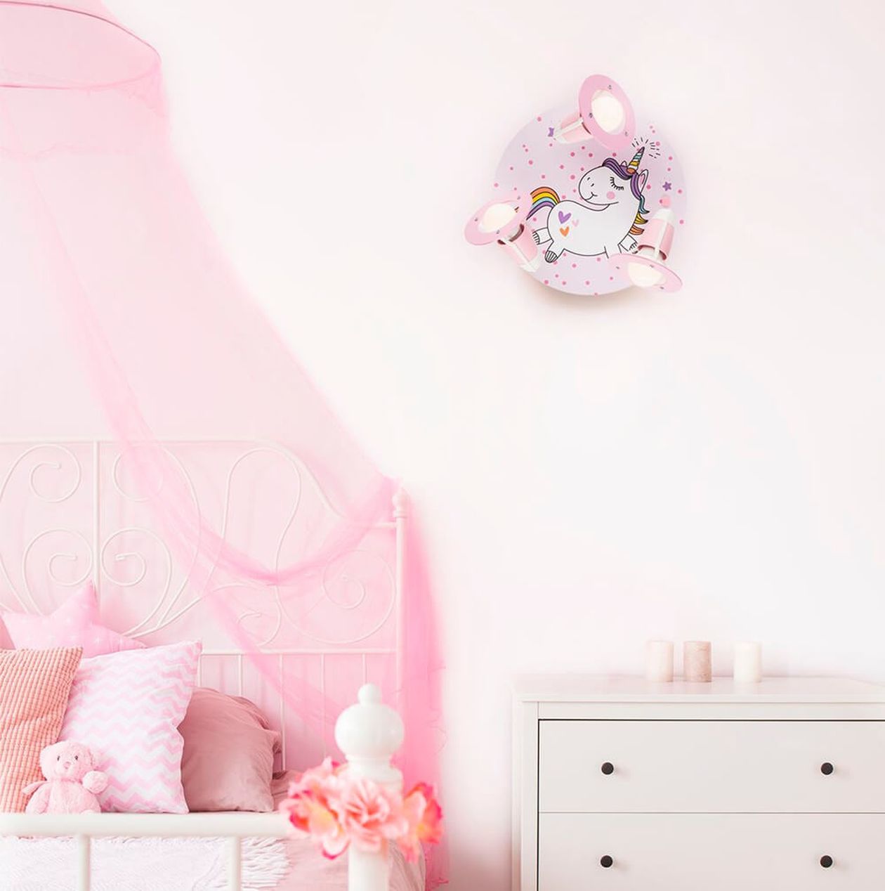 Rosa Mädchenzimmer mit Metallbett und rosa Baldachin. An der Wand eine rosa Kinderleuchte in Einhornform mit mehreren Spots. Daneben eine weiße Kommode mit Dekoartikeln.