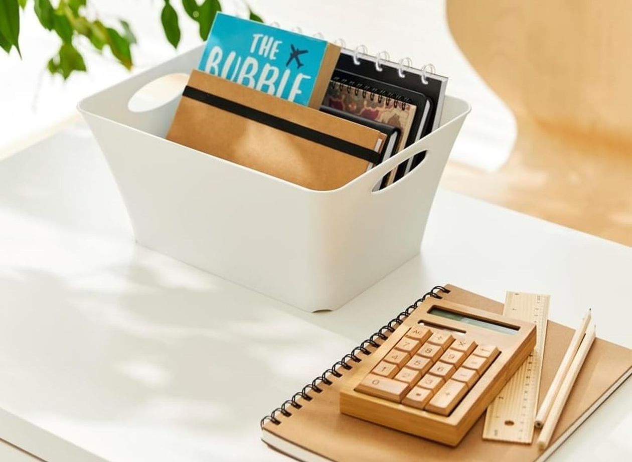 Weiße Aufbewahrungsbox mit seitlichen Grifföffnungen, gefüllt mit Notizbüchern und Heften, auf einem Schreibtisch neben einem Taschenrechner aus Holz.