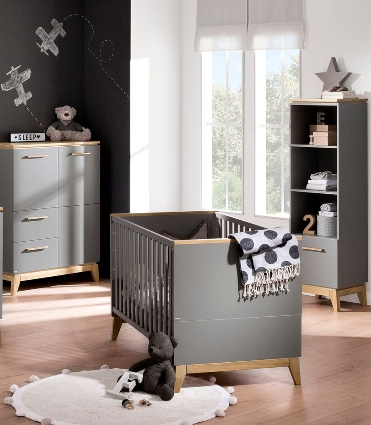 Vollständig eingerichtetes Babyzimmer mit grau-holzfarbenem Babybett, passender Wickelkommode und Schrank sowie schwarzer Tafelwand mit gezeichneten Flugzeugen im Hintergrund.
