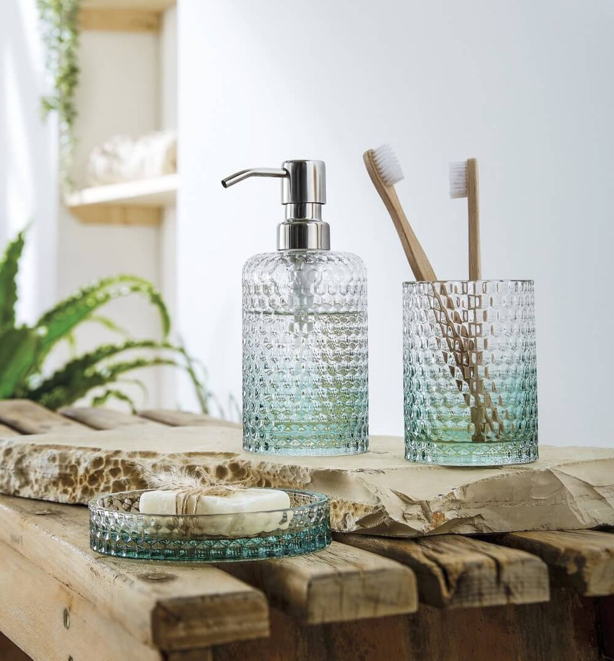 Strukturierter Glas-Seifenspender und Zahnbürstenhalter auf einer Holzoberfläche, mit einer Seifenschale und Pflanzen im Hintergrund.