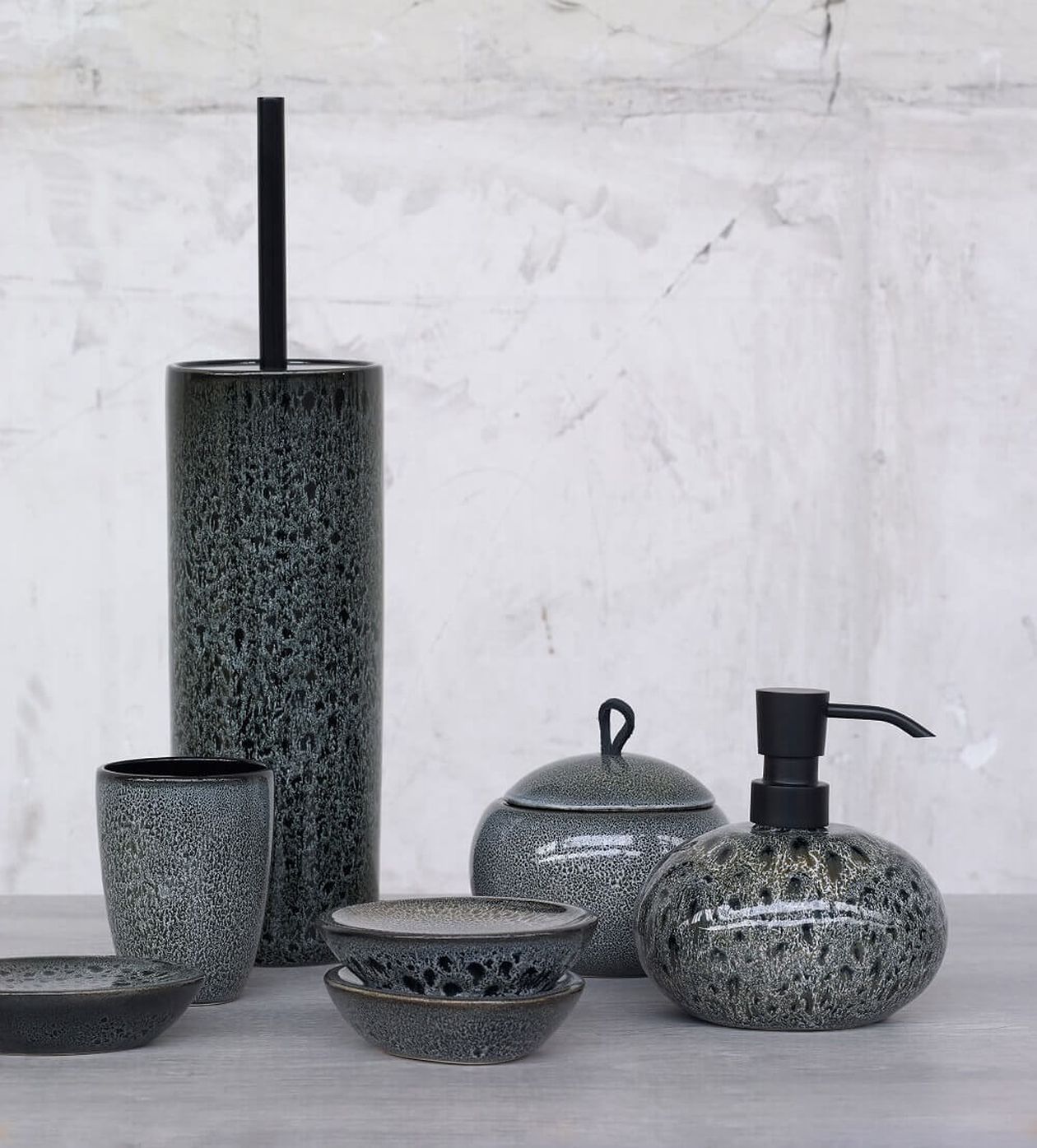 Set aus schwarzen Keramik-Badezimmeraccessoires mit strukturierter Oberfläche, einschließlich Seifenspender, Zahnputzbecher und behälter mit Deckel auf einer grauen Oberfläche.