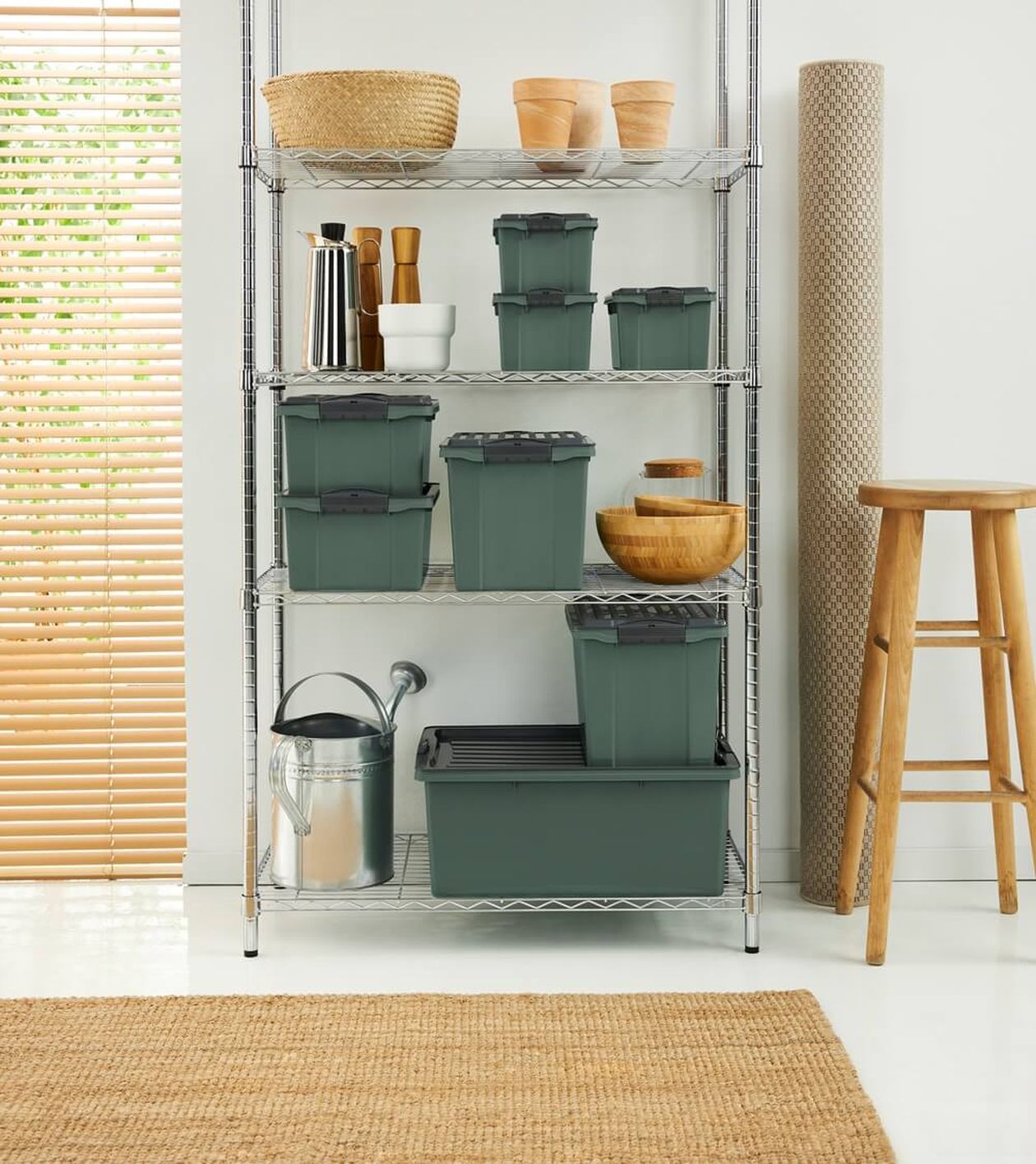 Metallregal mit grünen Vorratsbehältern, Holzschüsseln und Töpfen. Daneben eine Gießkanne und eine gerollte Matte. Rechts steht ein Hocker.