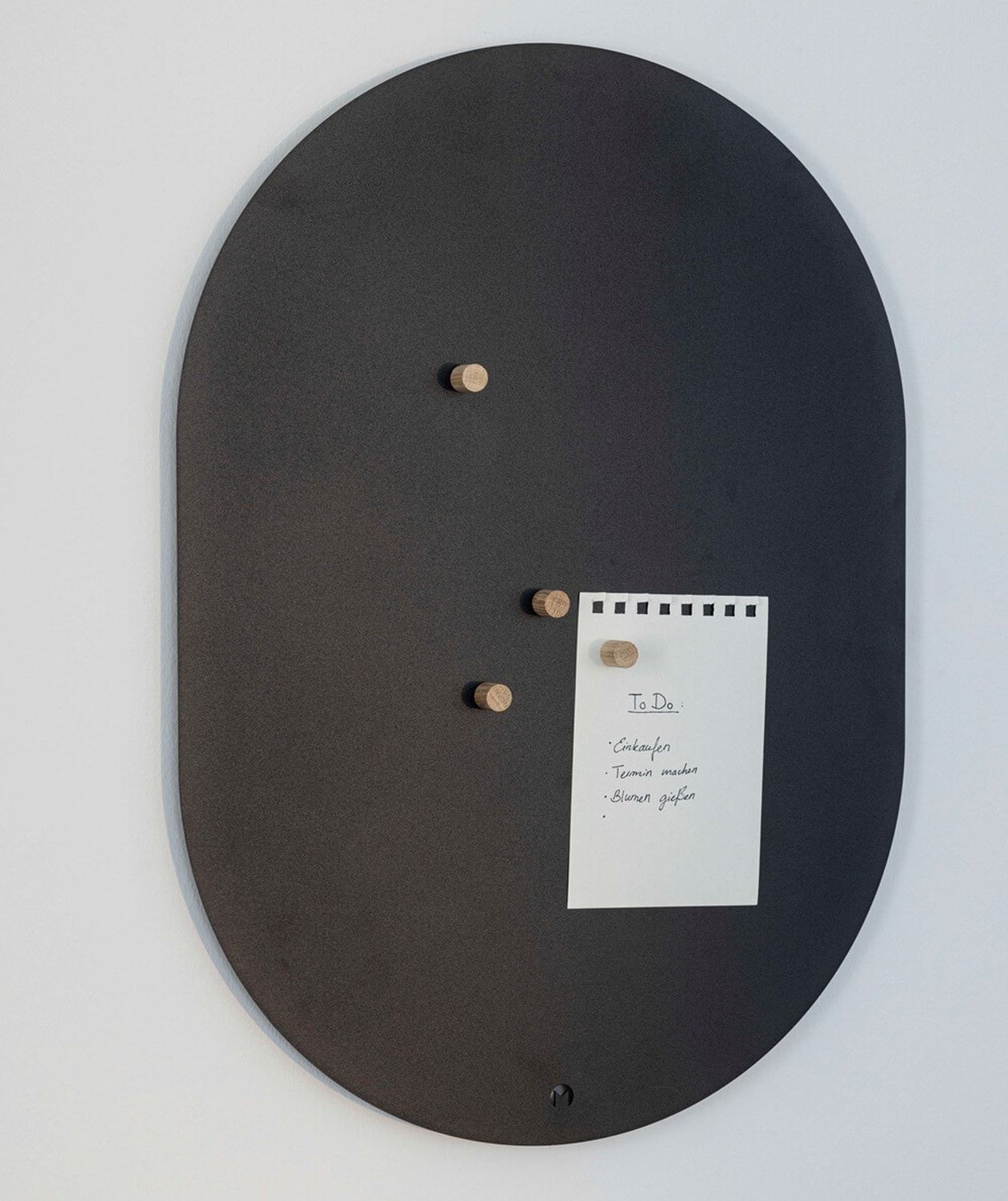 Ovale Magnetwand in Dunkelgrau mit kleinen runden Holzmagneten, an denen ein Notizzettel befestigt ist.