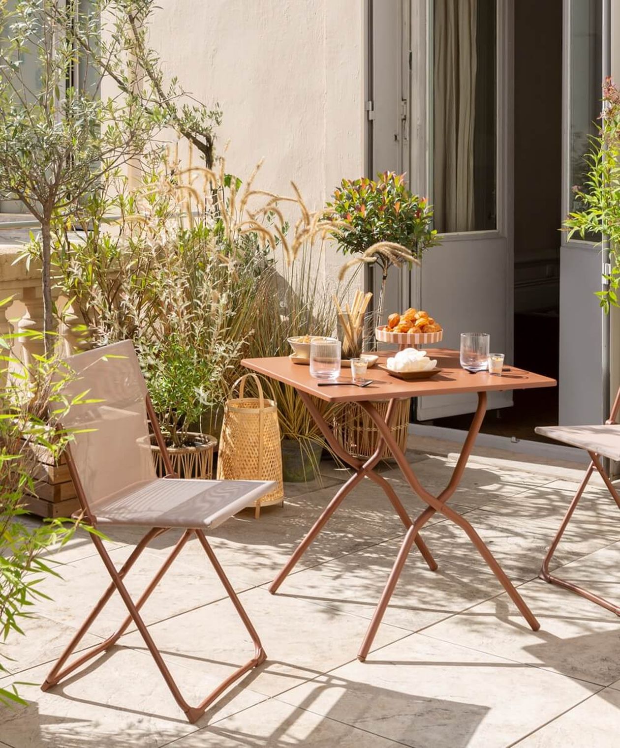 Zwei rosafarbene Klappstühle mit Textilbespannung und Metallgestell an einem passenden Klapptisch auf einer sonnigen Terrasse mit mediterranen Pflanzen.