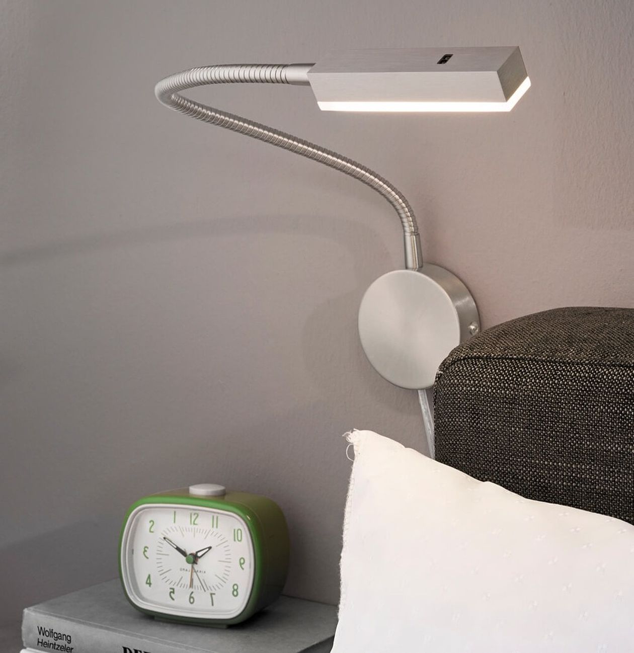 Silberne LED-Wandleuchte mit flexiblem Schwanenhals, montiert über einem Bett. Daneben ein grüner Retro-Wecker auf einem Bücherstapel.